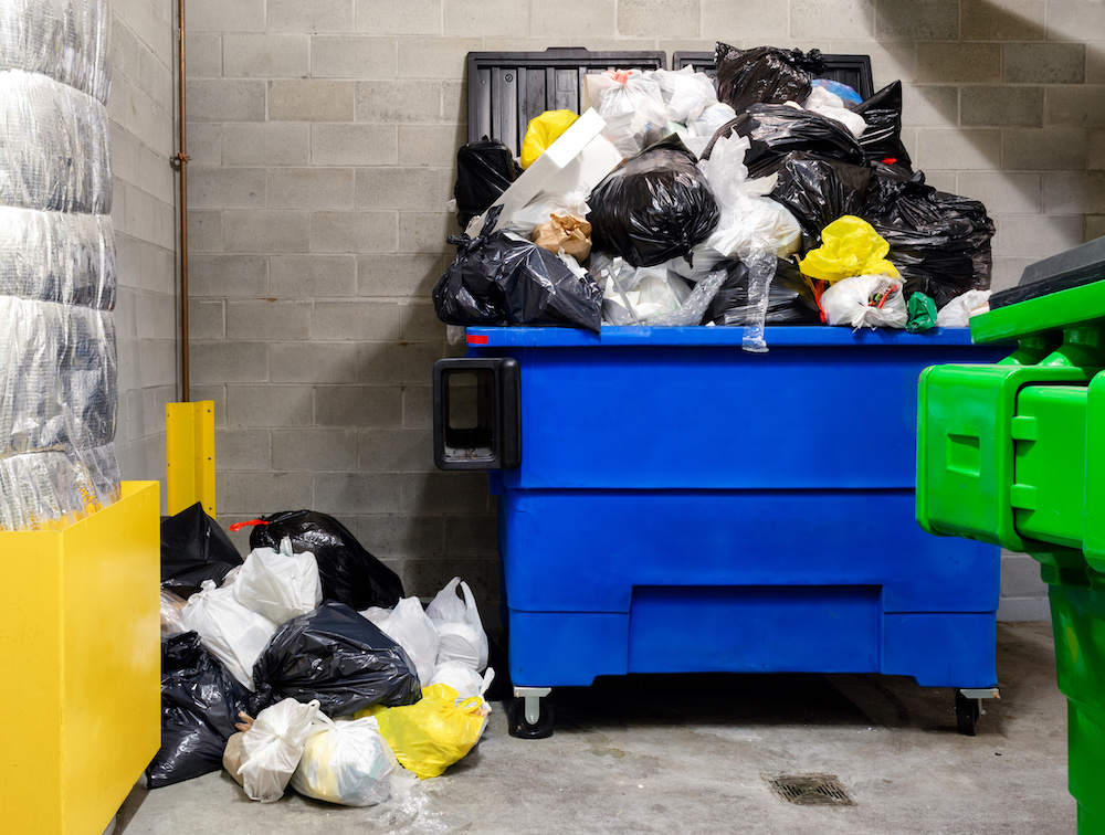 Overloaded household garbage container in garbage room of residential building. Waste management company has missed pickup due to snowstorm and people keep pilling it up. Indoors.