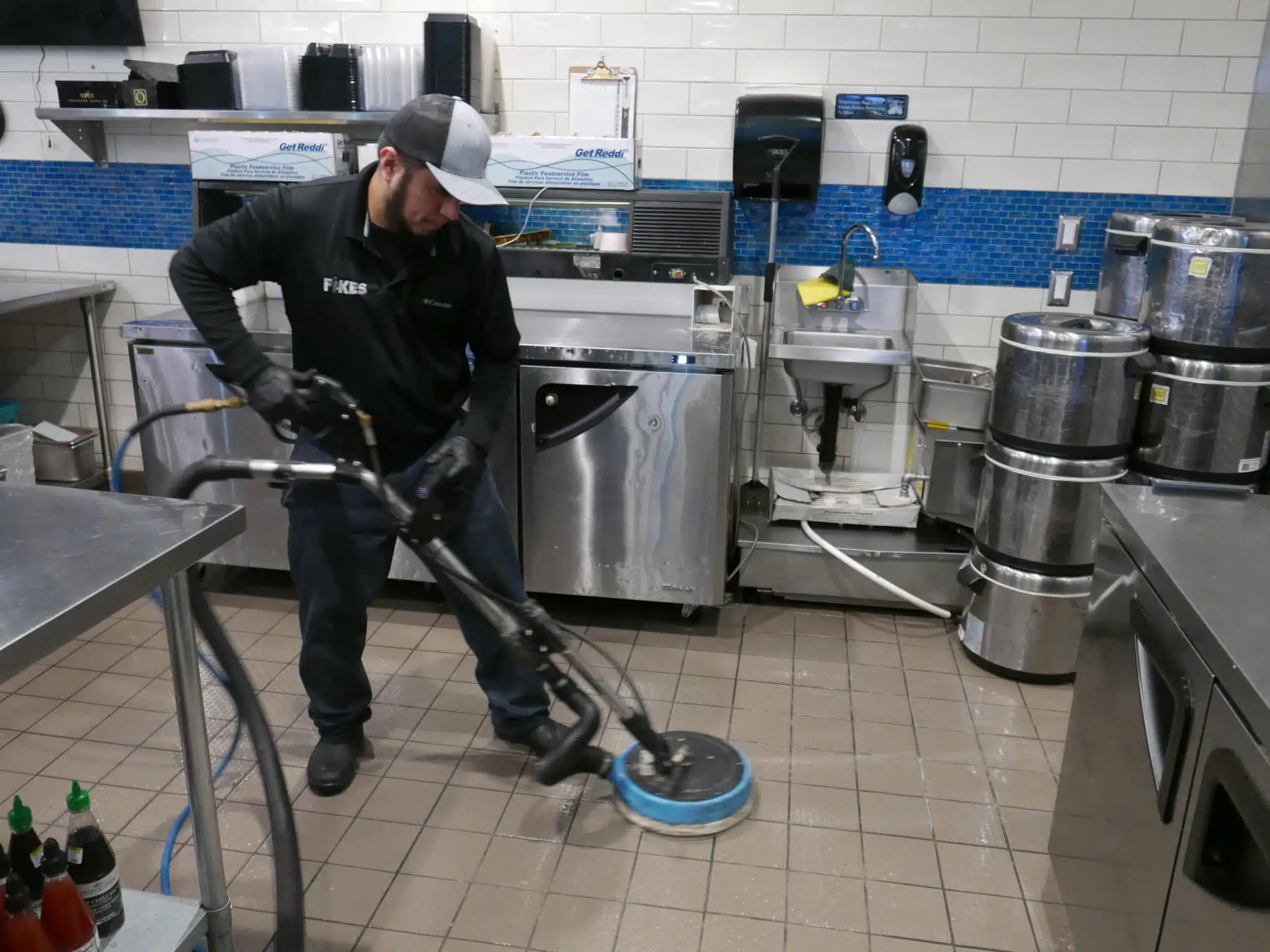 How High Summer Temperatures Fuel Commercial Kitchen Fruit Flies Man wearing a black uniform deep cleaning a commercial kitchen floor with a machine